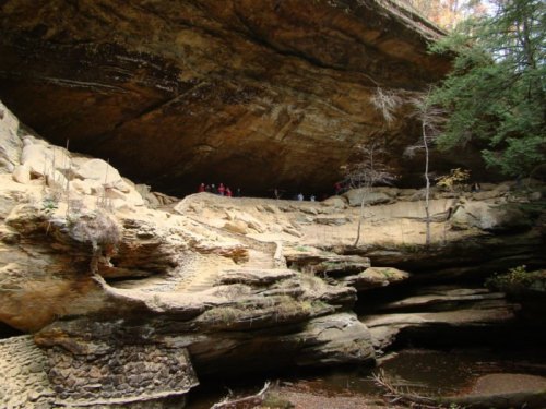 A rocky cliff with a large overhang, featuring trees and a group of people in the background near a stream.