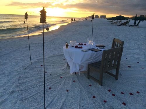 A romantic beach dinner setup at sunset, featuring a table with a white tablecloth, chairs, and rose petals on the sand.