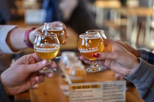Four hands clink small glasses of beer in a cozy brewery setting, with a menu visible in the background.