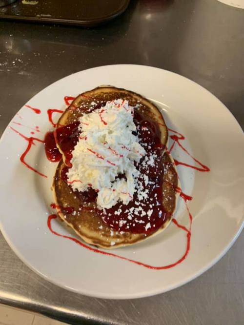 A plate of pancakes topped with whipped cream and strawberry sauce, drizzled with red syrup.