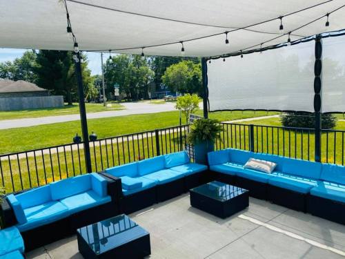 A cozy outdoor seating area with blue couches, a glass table, and string lights under a canopy, overlooking a grassy street.
