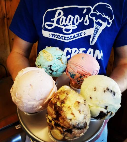 A person holds a tray with six colorful ice cream cones in various flavors, wearing a blue shirt that says "Lago's Homemade Ice Cream."