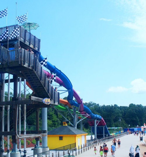 Colorful water slide at an amusement park, with people walking along a boardwalk under a clear blue sky.
