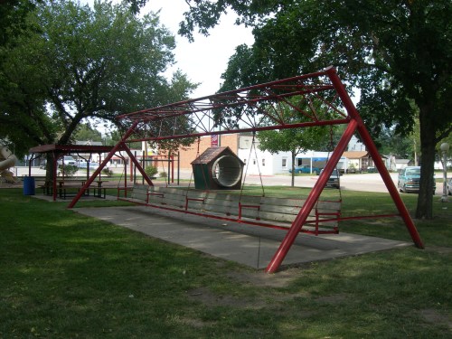 A red swing set with a tunnel slide in a grassy park, surrounded by trees and a few buildings in the background.