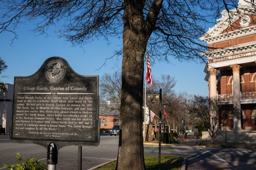 Historical marker for Oliver Hardy, with a tree and a building in the background, in a town square setting.
