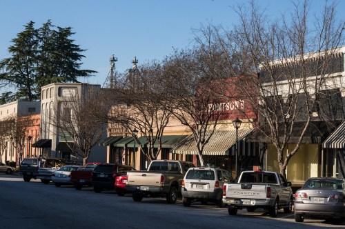 A quiet street lined with shops and parked cars, featuring bare trees and a clear blue sky.