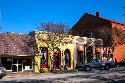Colorful storefronts line a street under a clear blue sky, with a mix of trees and parked vehicles nearby.