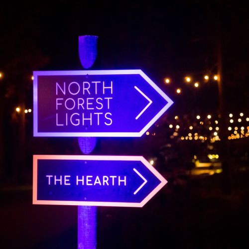 A sign at night with arrows pointing to "North Forest Lights" and "The Hearth," illuminated by colorful lights.
