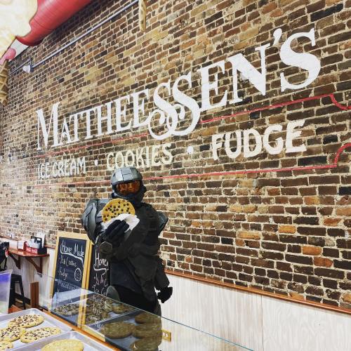 A person in a futuristic costume holds a large cookie in front of a brick wall with a sign for Mathiessen's ice cream shop.