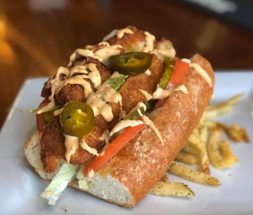 A crispy fried fish sandwich topped with jalapeños, tomatoes, lettuce, and drizzled with sauce, served with fries.