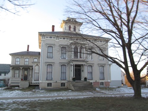 Victorian-style house with ornate architecture, large front steps, and a tree in the foreground, set in a snowy landscape.