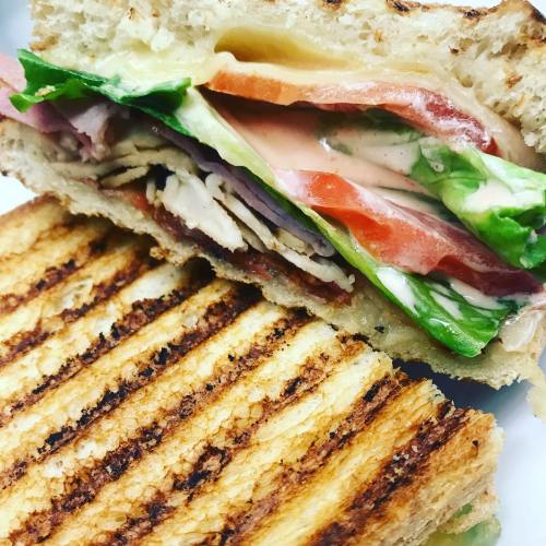 Close-up of a grilled sandwich with layers of turkey, ham, cheese, lettuce, and tomato, showcasing toasted bread.