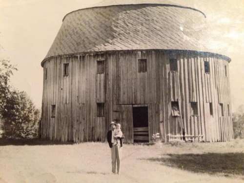 A man holds a child in front of a large, round wooden barn in a black-and-white photo.