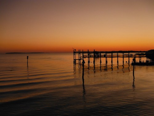 A serene sunset over calm waters, with a wooden pier reflecting in the water's surface.
