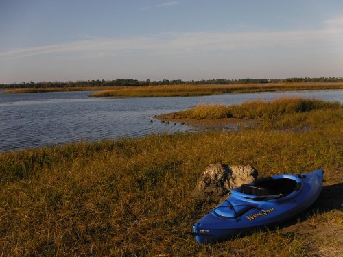 A blue kayak rests on the shore of a calm waterway, surrounded by tall grass and a clear blue sky.