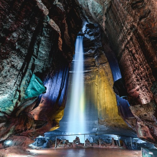A stunning cave with a tall waterfall cascading down colorful rock formations, illuminated by soft lights.