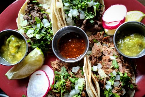 A vibrant plate of tacos with beef, cilantro, onions, lime, radishes, and three small bowls of salsa.