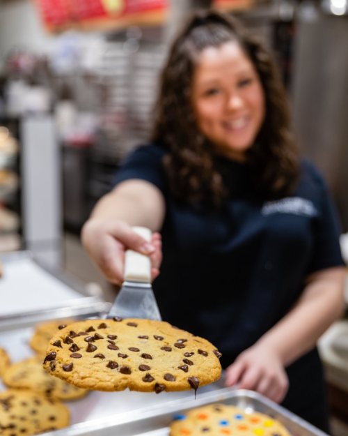 A smiling person holds a large chocolate chip cookie on a spatula, with more cookies on a tray in the background.