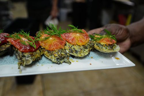 A plate of baked oysters topped with herbs and slices of citrus fruit, served in a kitchen setting.