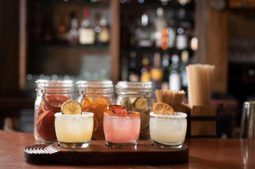 Three colorful cocktails with garnishes on a wooden tray, set against a blurred bar background with jars and bottles.