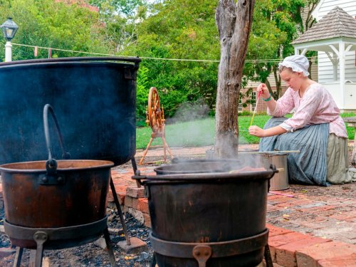 A woman in historical attire stirs a pot over an outdoor fire, surrounded by cooking pots and greenery.