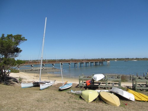 A serene waterfront scene with sailboats on the shore, a wooden pier, and clear blue skies.