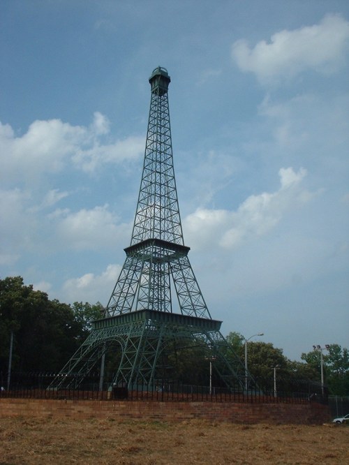 A replica of the Eiffel Tower stands tall against a cloudy sky, surrounded by trees and a fence.
