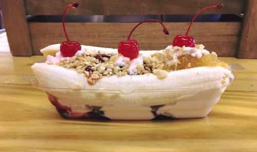 A banana split topped with whipped cream, nuts, and three cherries, served in a clear container on a wooden table.