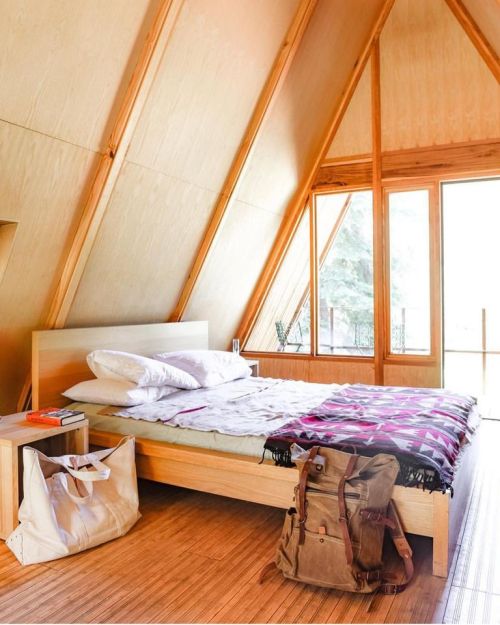 Cozy A-frame bedroom with a wooden bed, large windows, and a backpack on the floor, surrounded by natural light.