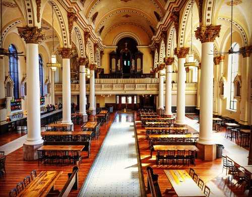 A spacious, ornate hall with tall columns, wooden tables, and a balcony, featuring intricate architectural details.