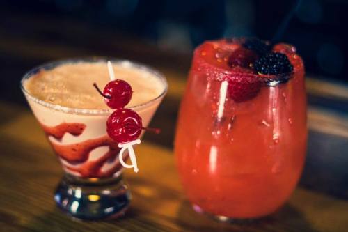Two colorful cocktails on a wooden bar: one with cherries and a swirl, the other garnished with berries.
