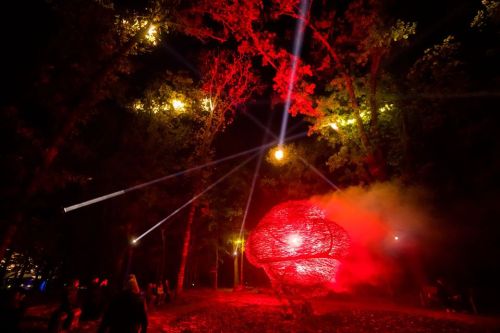 A glowing red sculpture surrounded by trees, illuminated by beams of light in a nighttime forest setting.