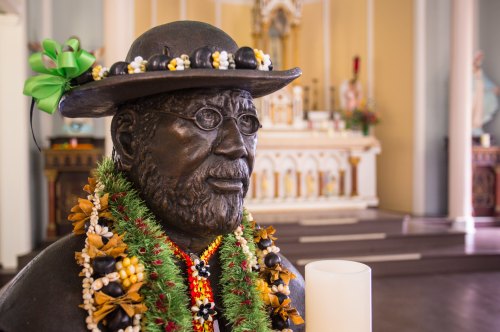 A bronze statue wearing glasses and a hat, adorned with floral garlands, inside a church setting.