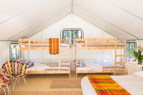 Interior of a cozy tent with two sets of wooden bunk beds, a colorful chair, and a warm blanket on a bed.