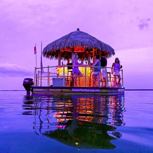 A floating tiki bar on calm water at sunset, with people enjoying the vibrant purple sky.