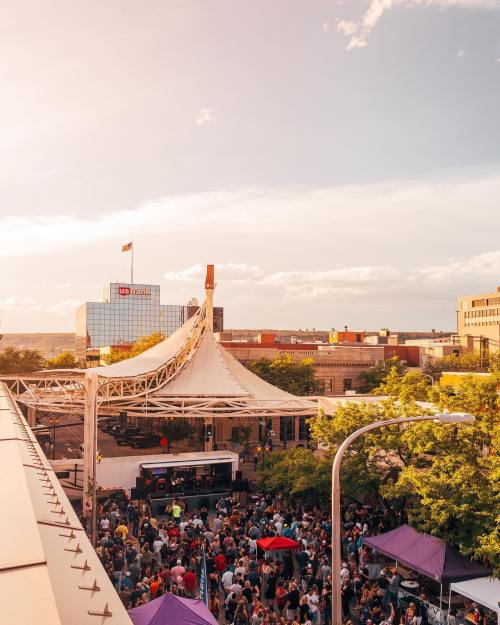A lively outdoor event with a large crowd under a canopy, surrounded by trees and buildings, during sunset.