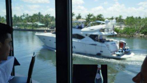 A view from inside a restaurant showing a yacht cruising on a calm waterway, with palm trees in the background.