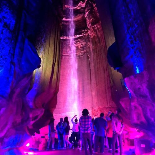 A group of people stands in a cave, illuminated by colorful lights, as a waterfall cascades down the rock walls.