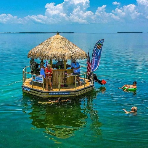 A floating tiki bar with people enjoying drinks, surrounded by clear blue water and a bright sky.
