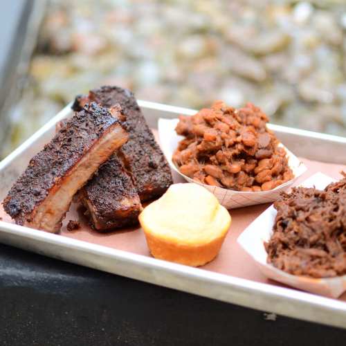 A tray with smoked ribs, baked beans, pulled pork, and a cornbread muffin, set against a blurred outdoor background.