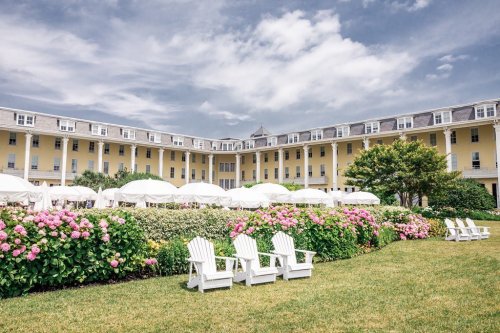 A sunny hotel courtyard with white chairs, blooming flowers, and umbrellas, set against a yellow building and cloudy sky.