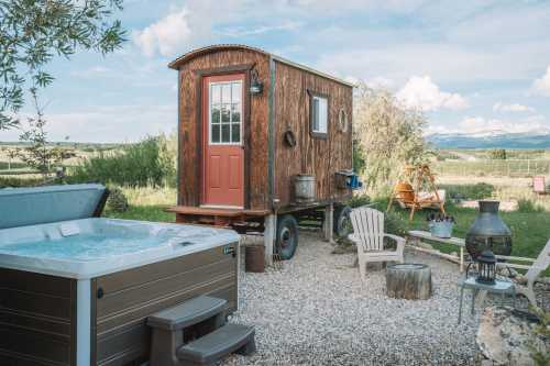 A cozy wooden tiny house on wheels beside a hot tub, surrounded by greenery and mountains in the background.