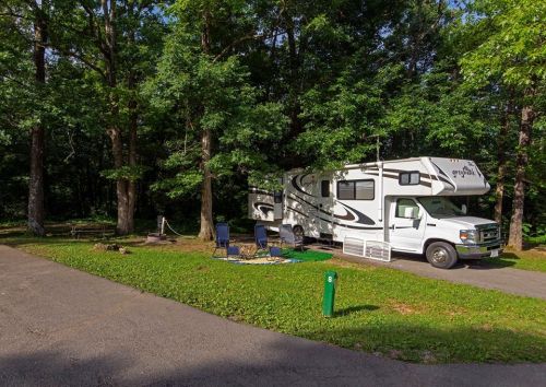 An RV parked in a wooded campsite, surrounded by green grass and trees, with chairs set up nearby.