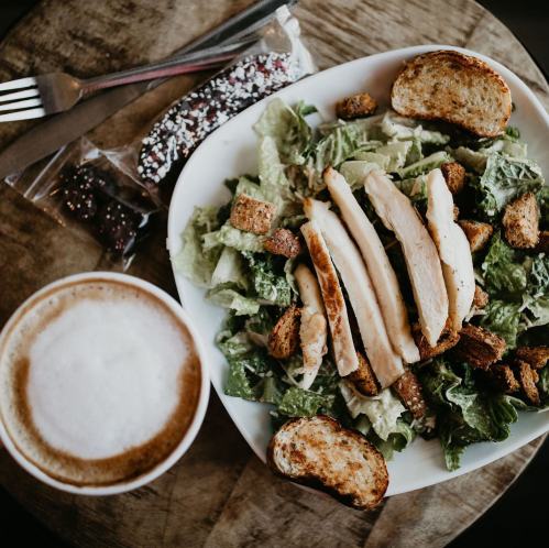 A bowl of Caesar salad with grilled chicken, croutons, and toasted bread, alongside a cup of cappuccino and snacks.