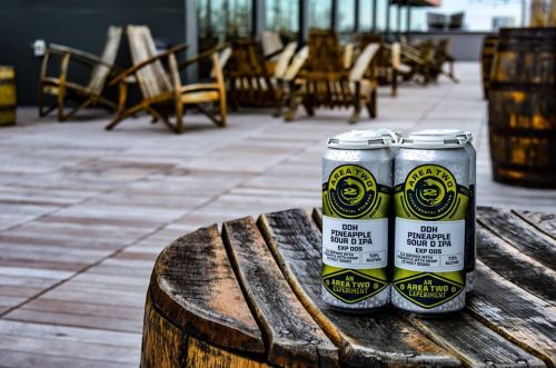 Two cans of Area Two's DDH Pineapple Sour IPA on a wooden table, with outdoor seating and barrels in the background.