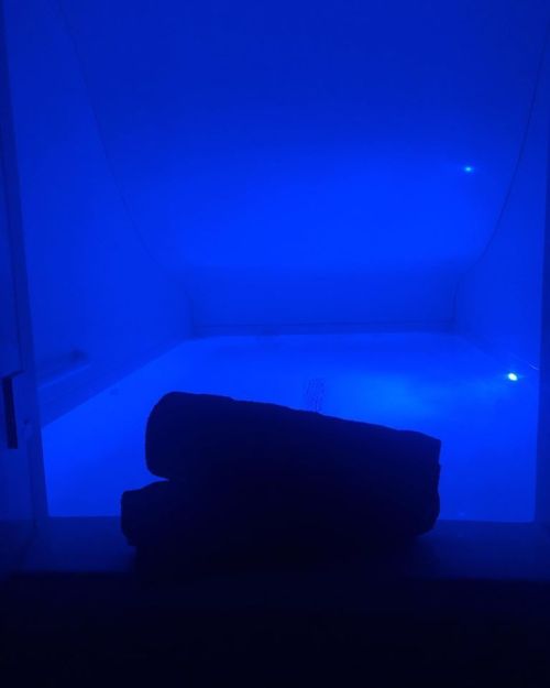 A dimly lit room with a blue-tinted float tank and two rolled towels in the foreground.