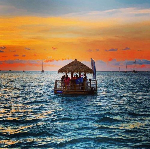 A thatched-roof hut on a floating platform in the ocean at sunset, with colorful skies and distant sailboats.