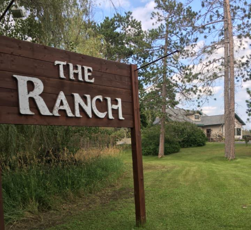 A wooden sign reading "The Ranch" stands near a grassy path leading to a house surrounded by trees.