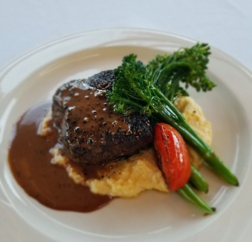 A plated dish featuring a steak with sauce, creamy polenta, roasted tomato, and broccolini on a white background.