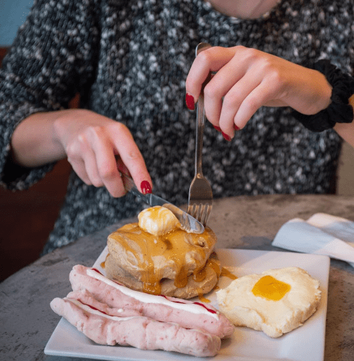 A person cuts into a pancake topped with butter and syrup, alongside sausage and a dollop of egg on a plate.